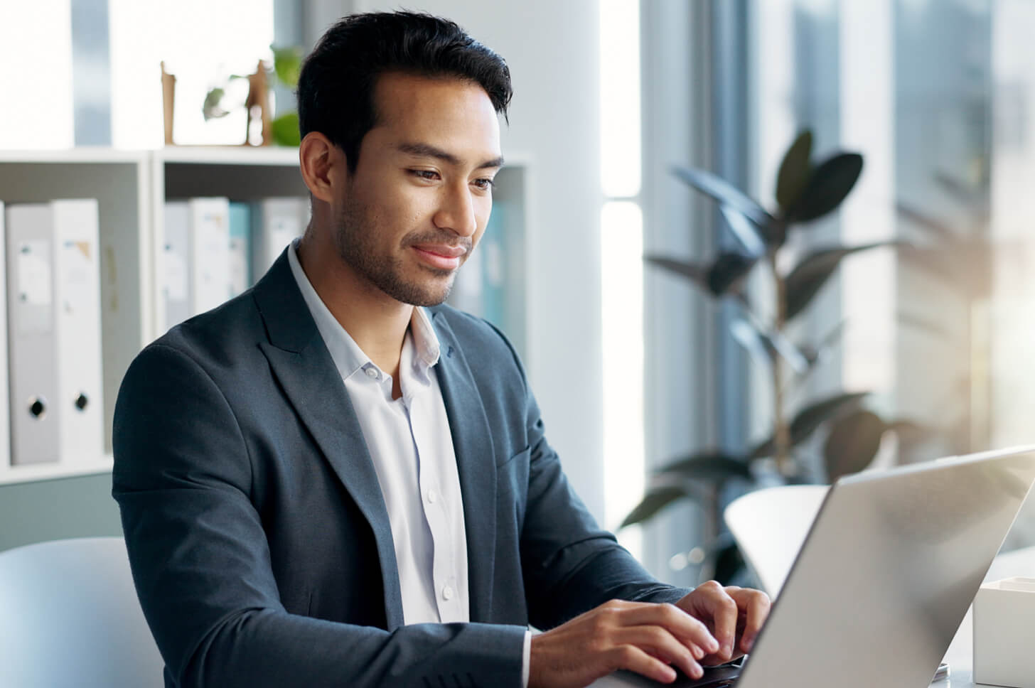 Man in a suit with a slight beard who is smiling while working at a laptop